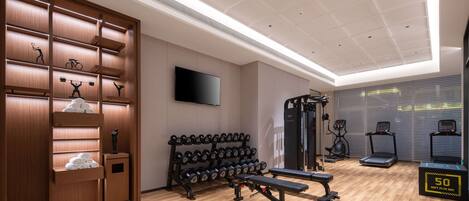 Un gimnasio moderno con suelo de madera, estantes para pesas, mancuernas, una prensa de banca, cintas de correr y una televisión montada en la pared.