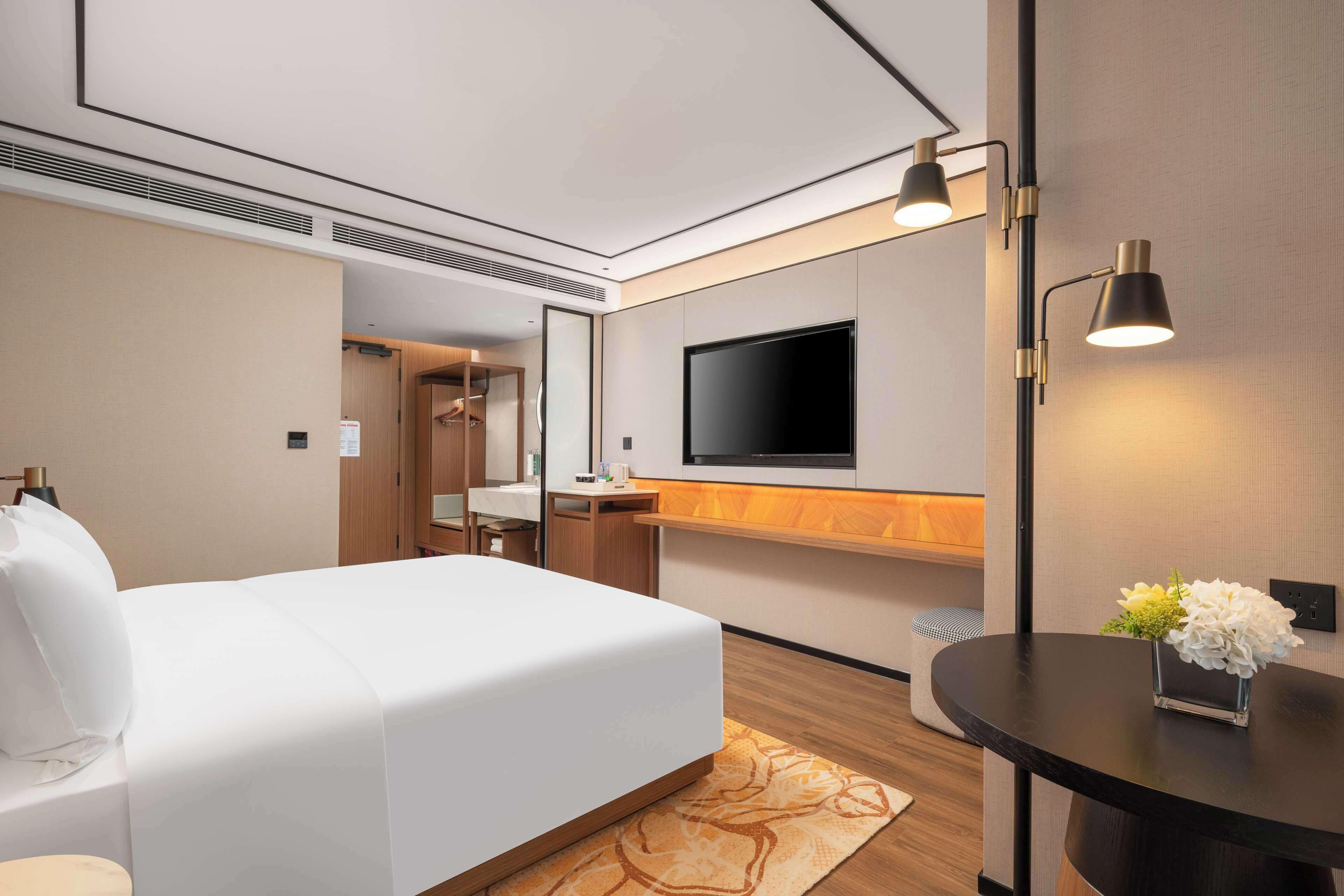 A hotel room with a large bed, a flat-screen TV mounted on the wall, a small table with a vase of flowers, and a desk with a chair.