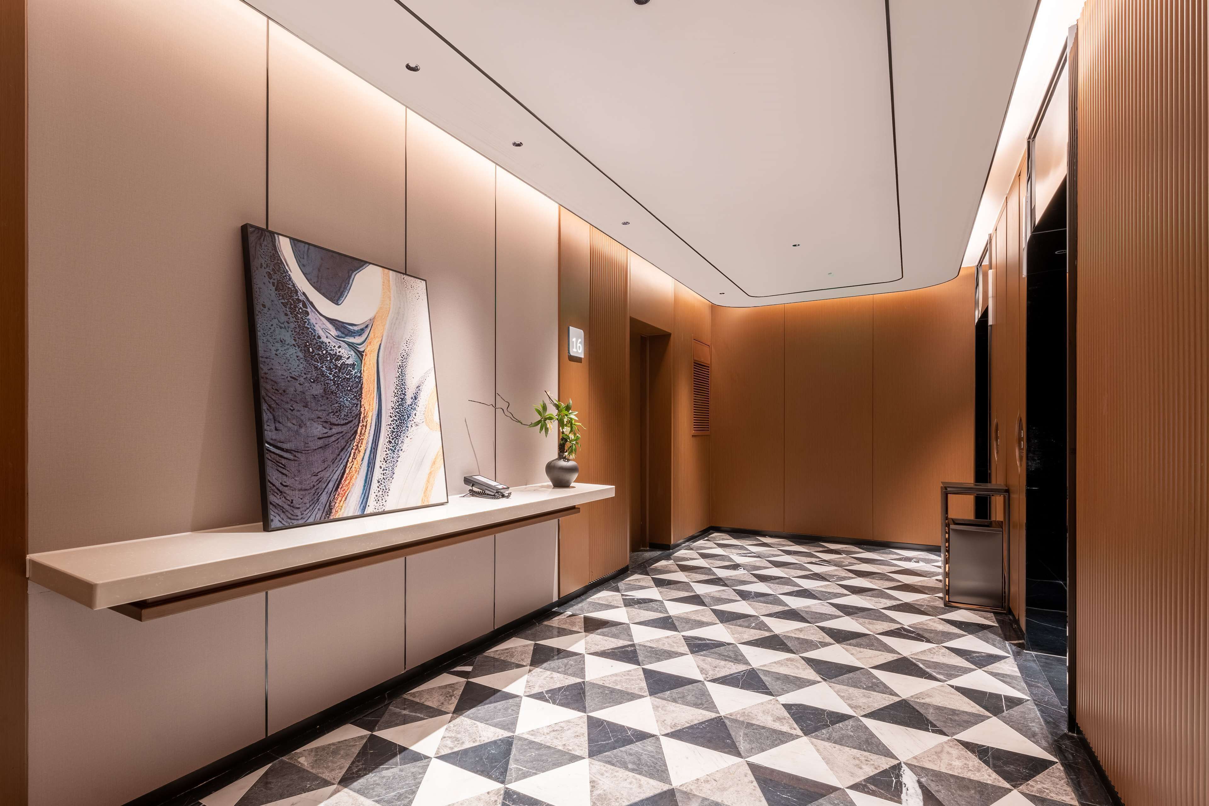 A modern hotel lobby with a geometric patterned floor, a reception desk, a large abstract painting, and a potted plant.