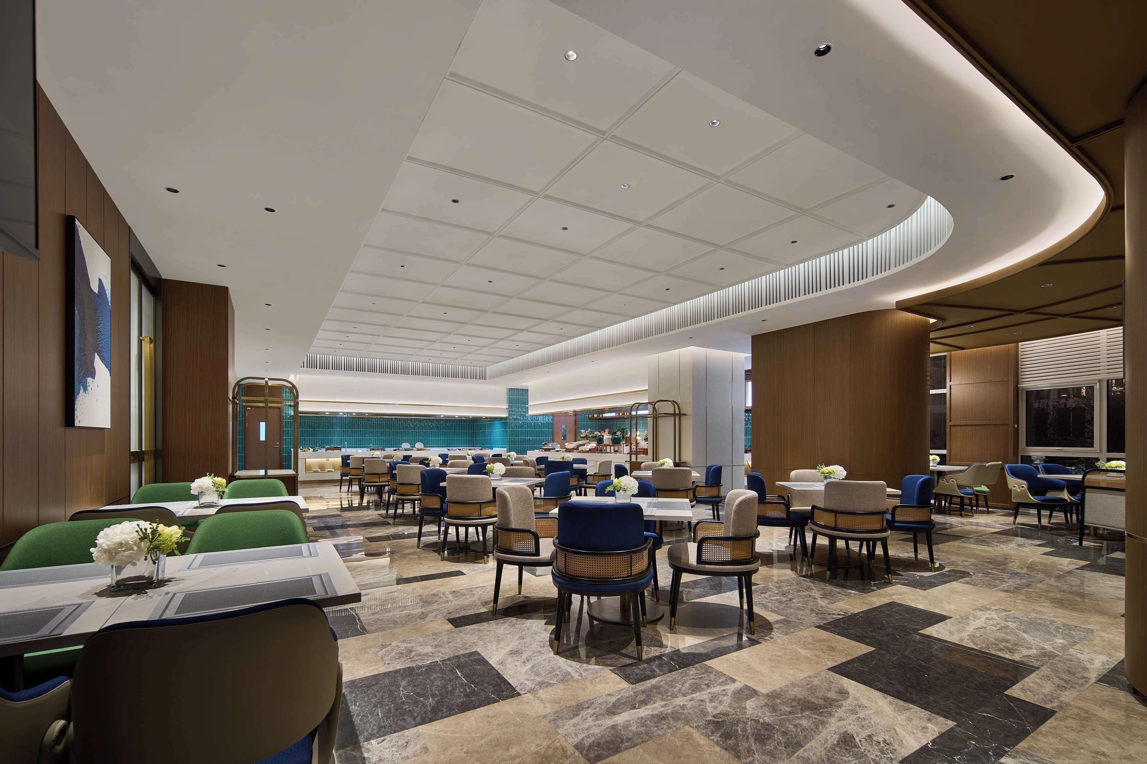 A modern hotel lobby with a spacious layout, featuring multiple dining areas with tables and chairs, a central seating area, and a large, curved ceiling design.