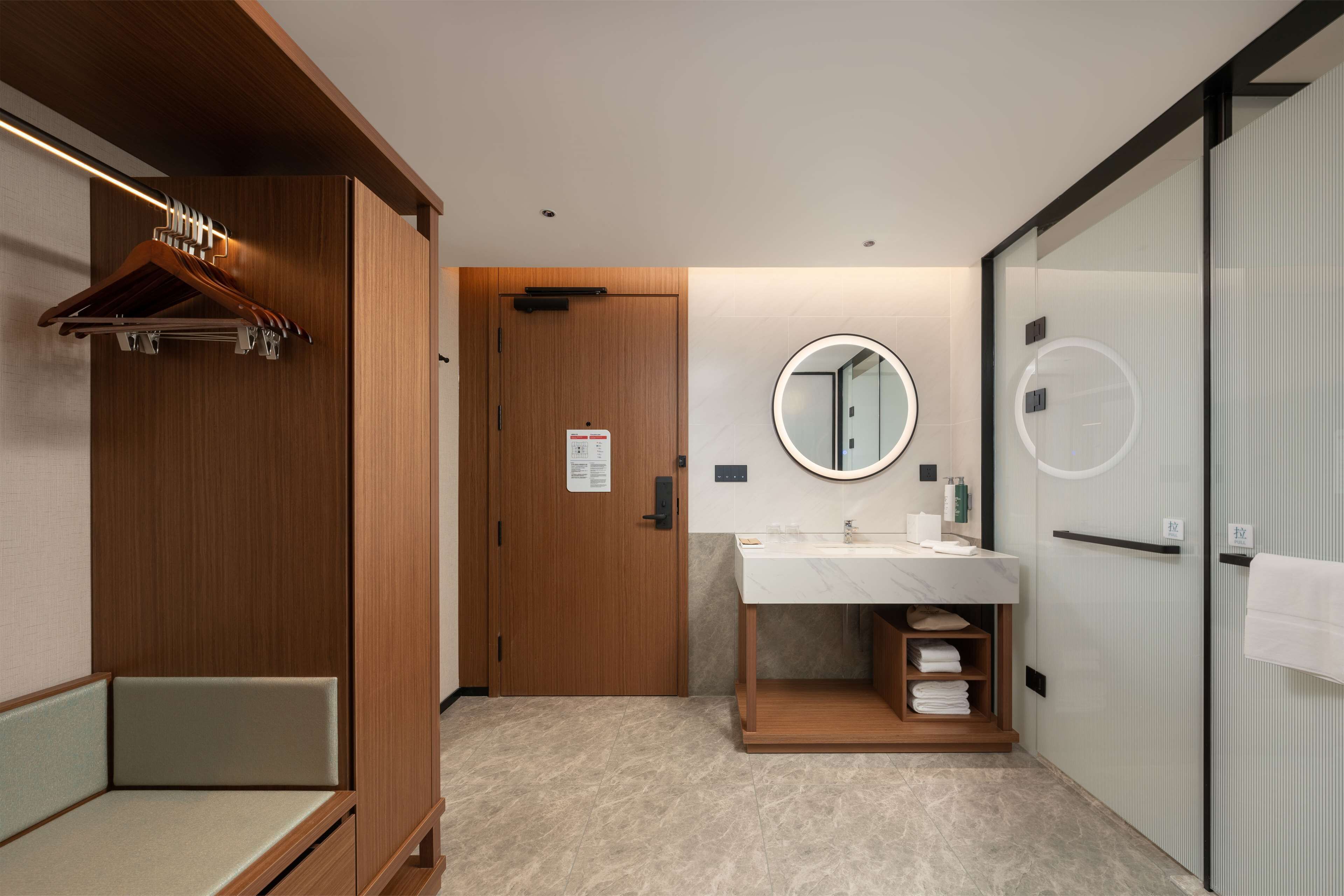 A modern bathroom with a wooden vanity, a round mirror, a towel rack, and a bench.