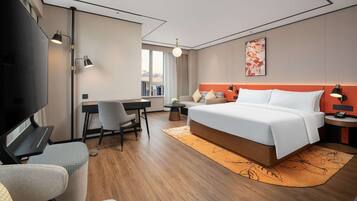 A modern hotel room with a large bed, a desk, a chair, a television, and a view of the city.