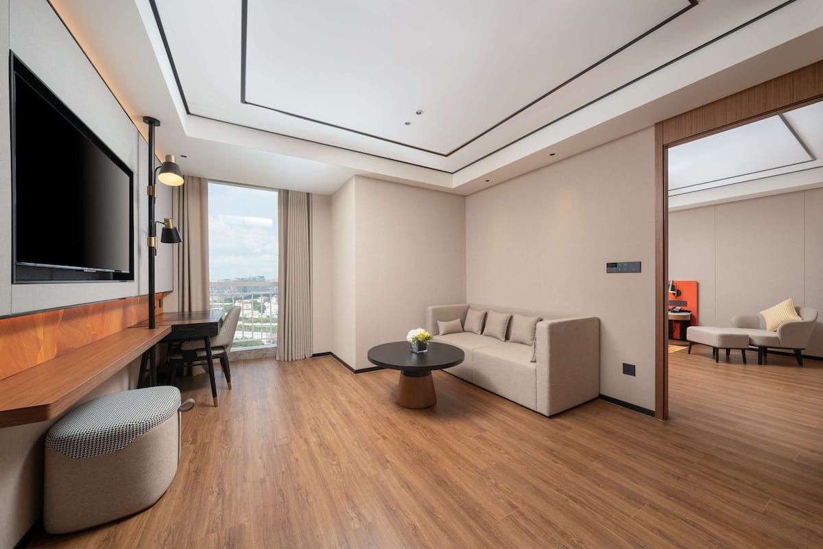 Ein modernes Hotelzimmer mit Holzboden, einer Couch, einem runden Couchtisch, einem an der Wand montierten Fernseher und einem großen Fenster mit Blick auf die Stadt.