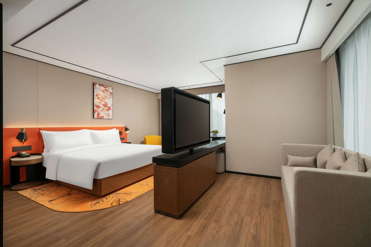 Ein modernes Hotelzimmer mit Bett, Fernseher auf einem Holzboden, einer Couch und einem kleinen Tisch mit Lampe.