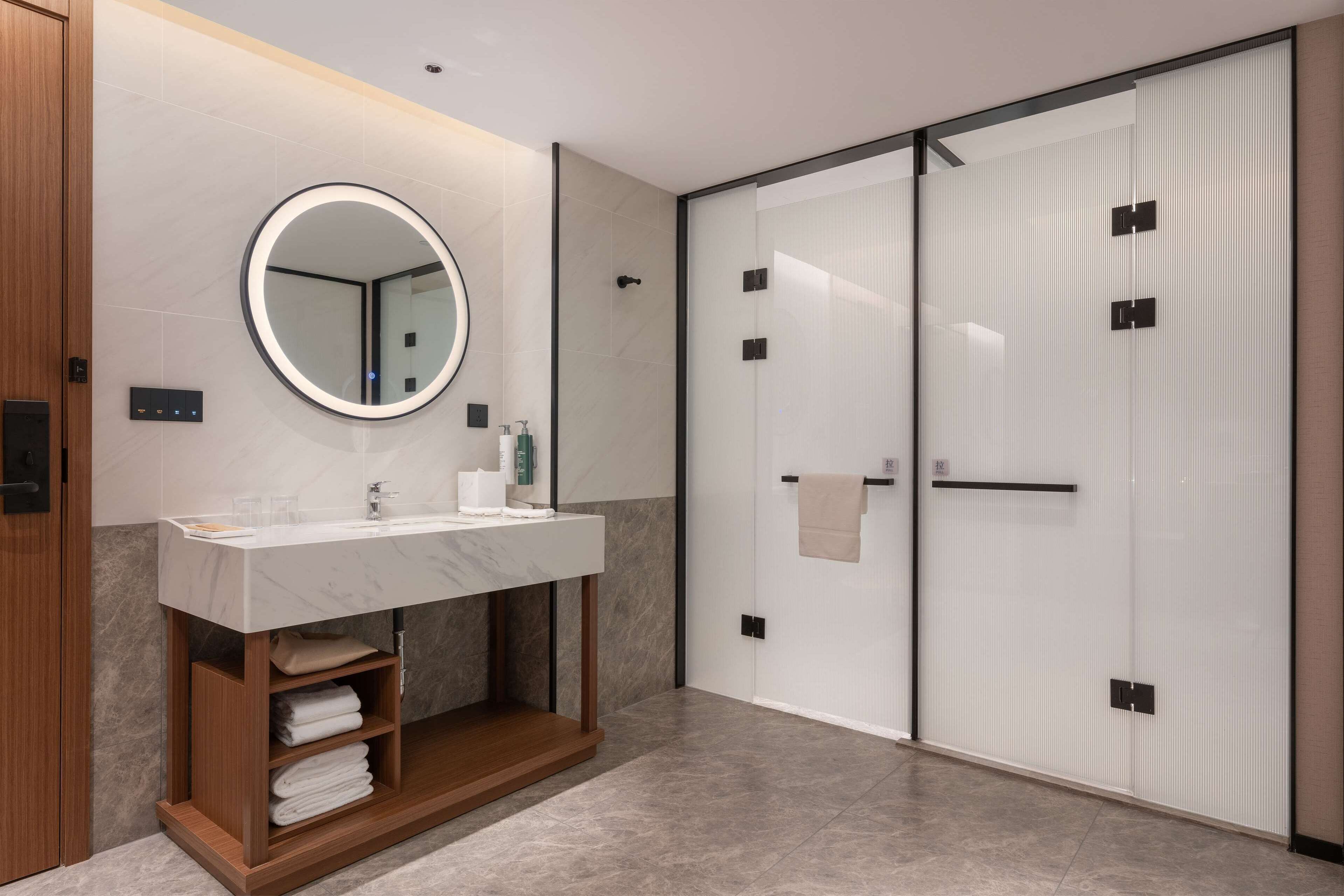 A modern bathroom with a marble sink, a round mirror, a wooden vanity, and a glass shower enclosure.