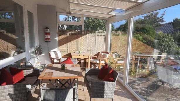 Een zonnig terras met rieten meubels, een houten tafel en uitzicht op de tuin door grote ramen.