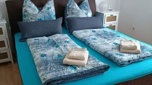 Een tweepersoonsbed met een blauwe spreidek en bijpassende kussens, een nachtkastje met een lamp, en een kunstwerk aan de muur.