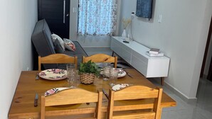 A dining area with a wooden table set for four, a dark door, a window with floral curtains, and a white cabinet with a flat-screen TV. - Casinha33, O Aconchego da Tríplice Fronteira! (Jardim Cataratas Foz do Iguaçu)