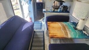 A compact RV interior with two blue cushioned seating areas, a wooden table with a wave design, and a kitchenette with a stove and sink. - The Holy Mackerel 
Family and pet friendly  (Perry)