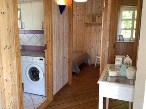Habitació senzilla amb rentadora, llit, taula petita amb objectes decoratius i finestra que dona a zona verda.