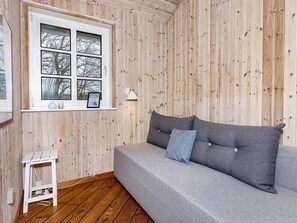 A room with a wooden paneled wall, a grey sofa with cushions, a small white table, and a window with a view of bare trees. - 4 Star Holiday Home in Thisted (Thisted)