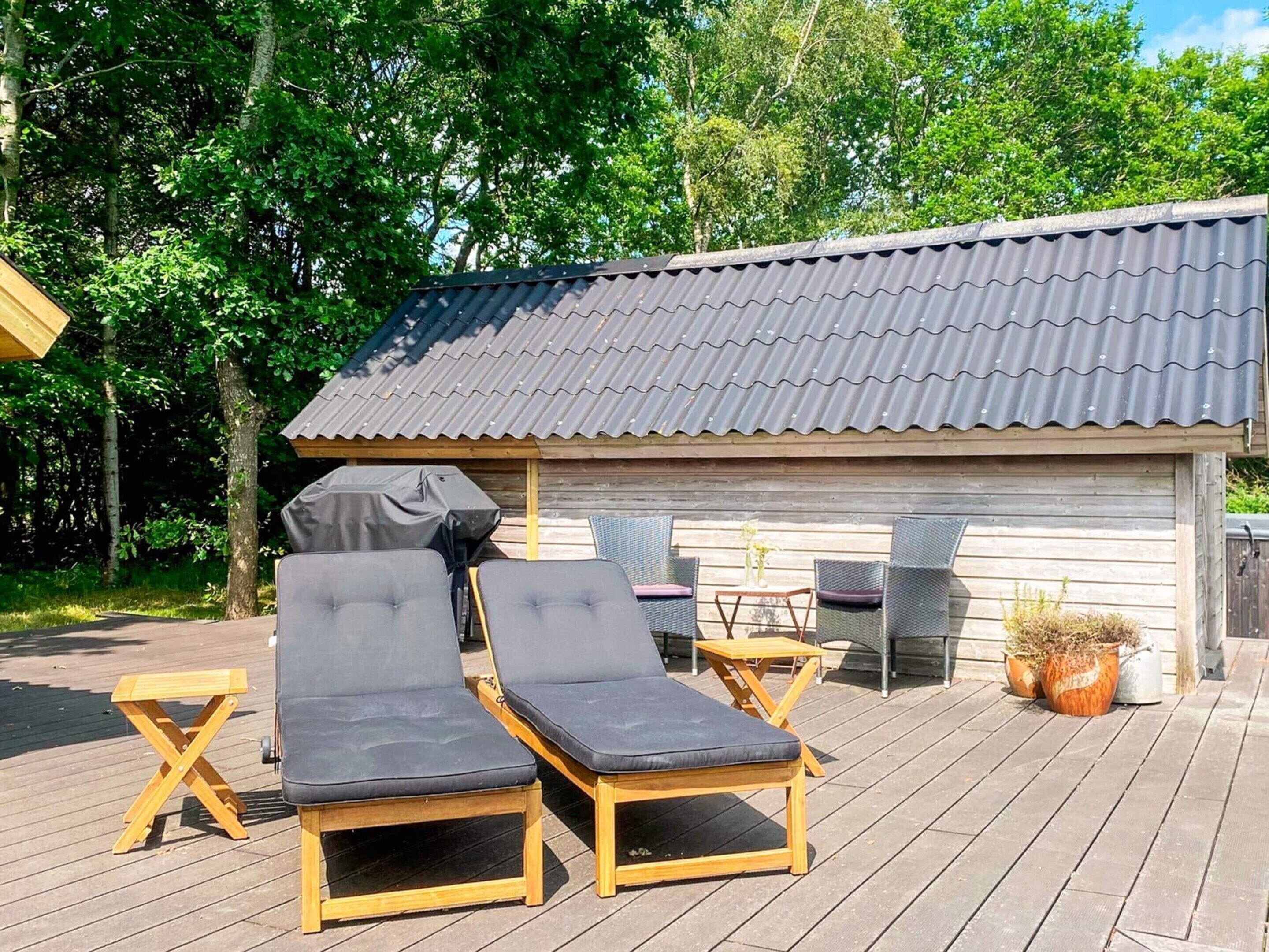 Une terrasse en bois avec deux chaises longues et une table, un barbecue couvert et un petit bâtiment doté d’un toit en tôle ondulée.