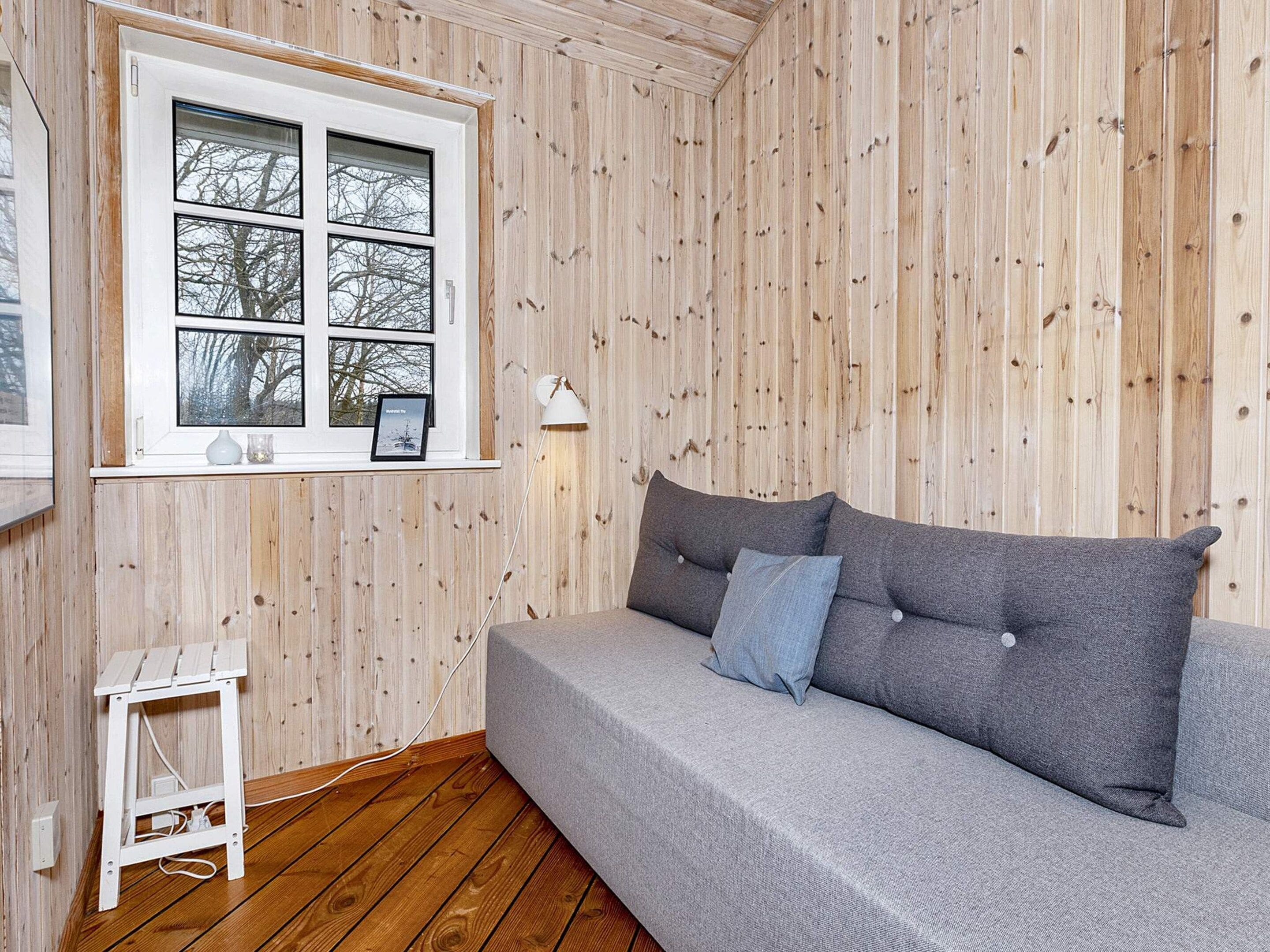 Una habitación con un revestimiento de madera, un sofá gris con cojines, una mesita blanca y una ventana con un marco de foto y una lámpara.