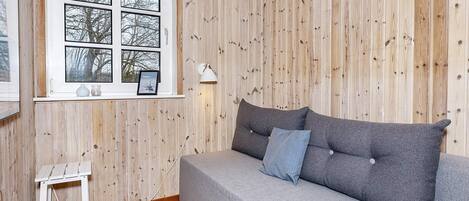 Una habitación con un revestimiento de madera, un sofá gris con cojines, una mesita blanca y una ventana con un marco de foto y una lámpara.