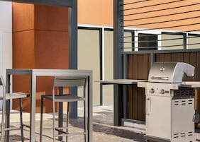 Un patio al aire libre con una mesa y sillas de metal, una parrilla y un edificio moderno con un muro de listones de madera al fondo.
