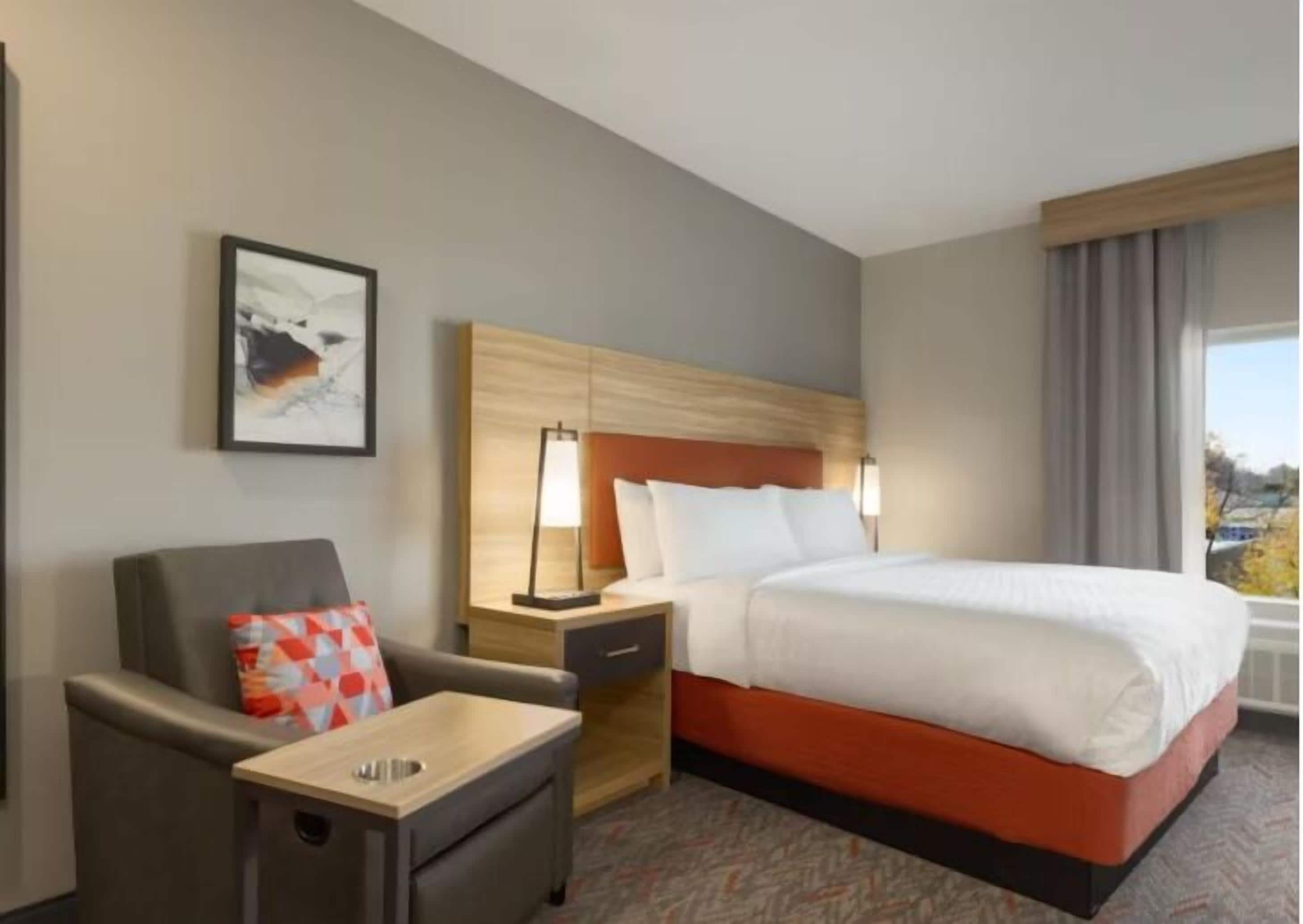 Habitación de hotel con cama, escritorio, silla, mesita de noche, lámpara y un cuadro en la pared.