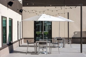 An outdoor seating area with tables and chairs under a large umbrella, string lights overhead, and a building entrance in the background. - EVEN Hotel Bozeman Yellowstone Intl Arpt​ by IHG (Belgrade)
