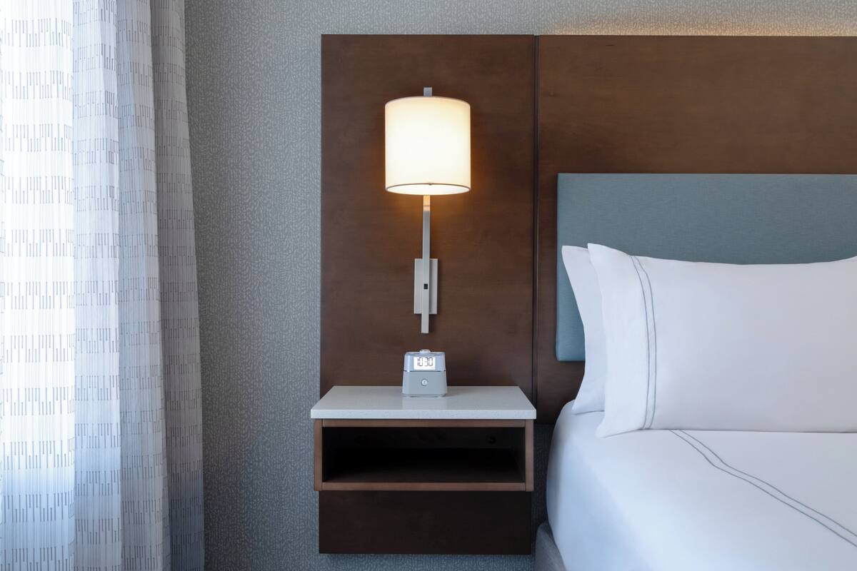 A neatly made hotel room with a bed, a nightstand with a lamp, and a digital clock.