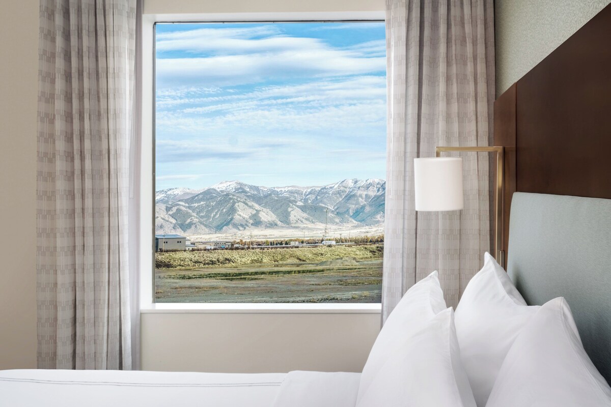 A hotel room with a large window offering a mountain view, a bed with white linens, and a bedside table with a lamp.