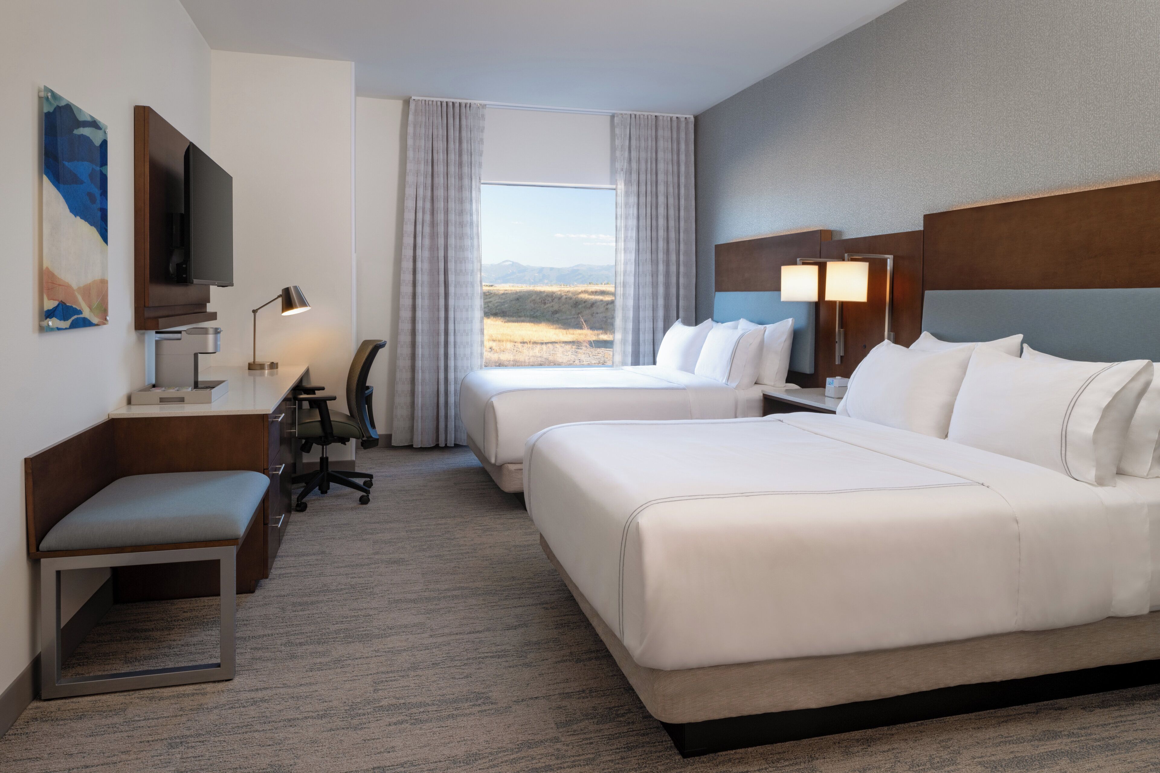 A hotel room with two beds, a desk with a chair, a TV, and a large window with a view of the landscape.