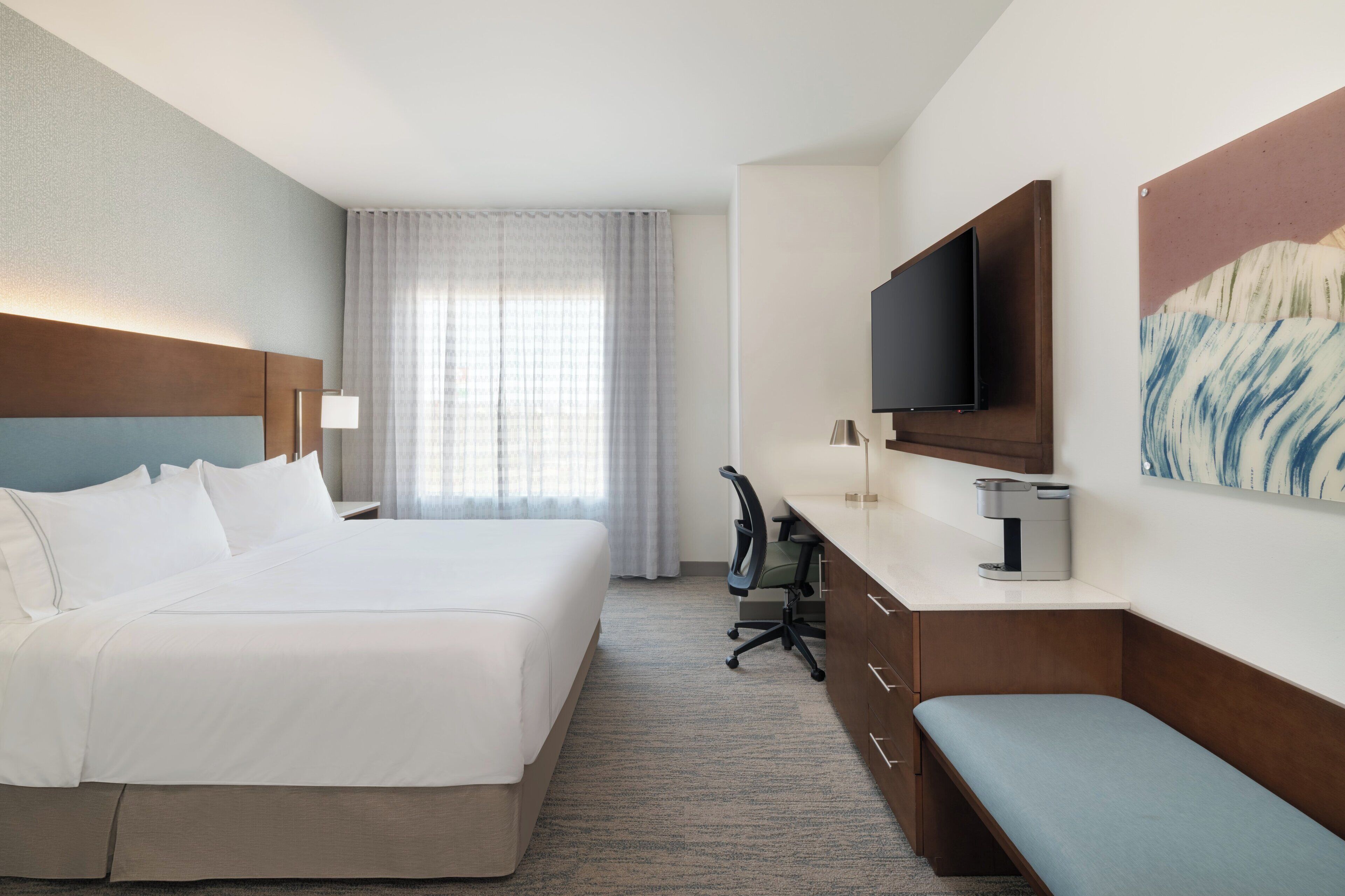 A hotel room with a large bed, a desk with a chair, a coffee maker, a TV, and a painting on the wall.