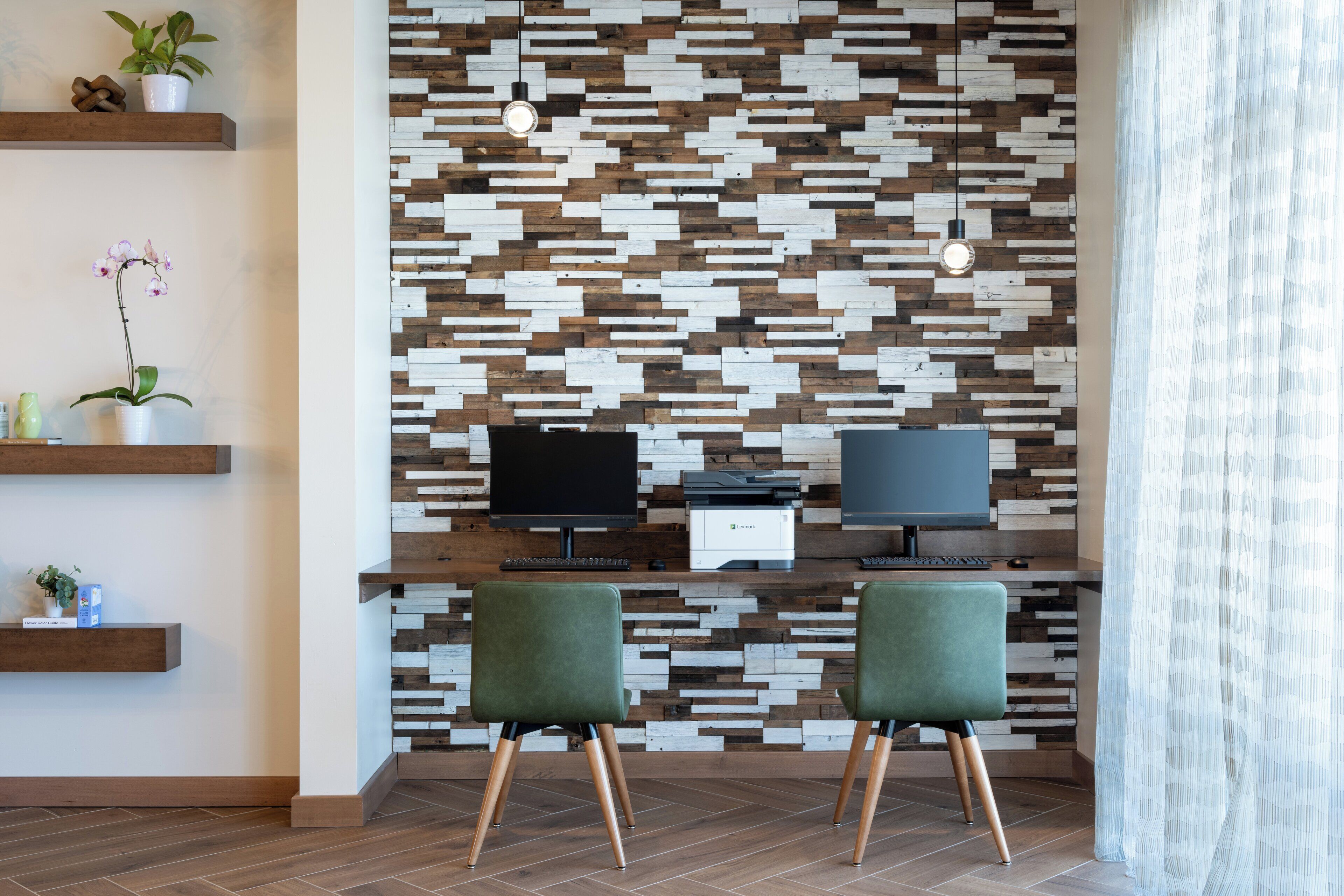 A modern office space with two desks, green chairs, a patterned wall, and shelves with plants.