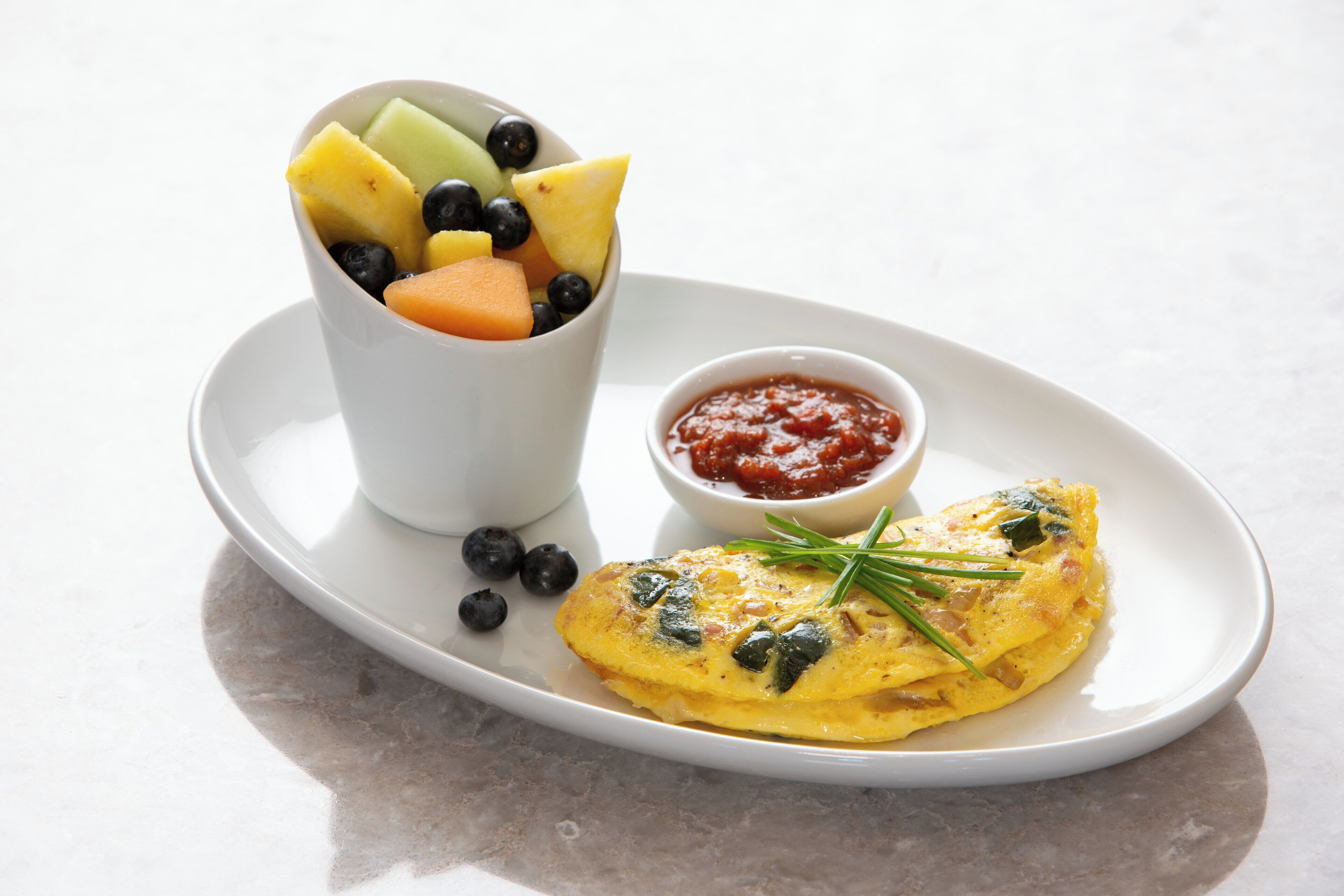 A plate with an omelette, a bowl of mixed fruit, and a small bowl of sauce.