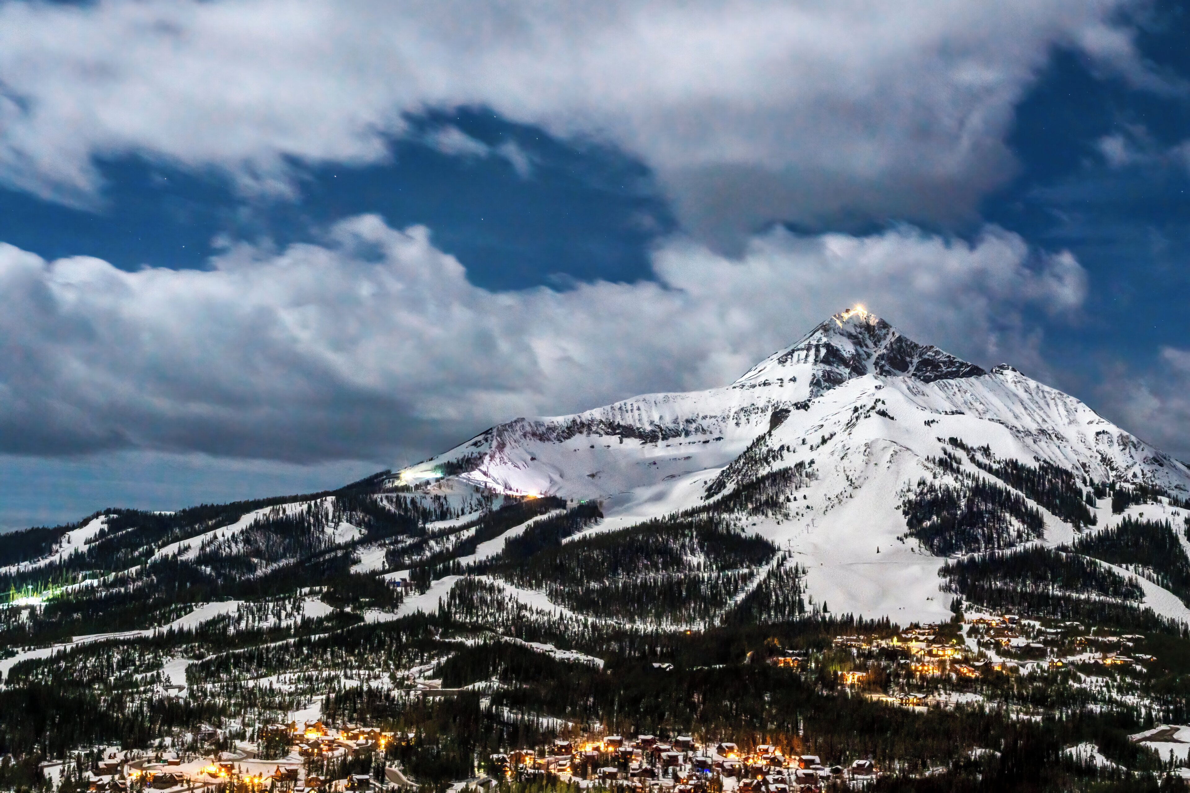 A snow-covered mountain with a town at its base, featuring illuminated buildings and a prominent peak.