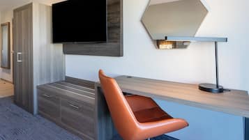 Una habitación de hotel moderna con un televisor de pantalla plana montado en un panel de madera, un escritorio con una computadora portátil, una silla naranja, un espejo y un armario.