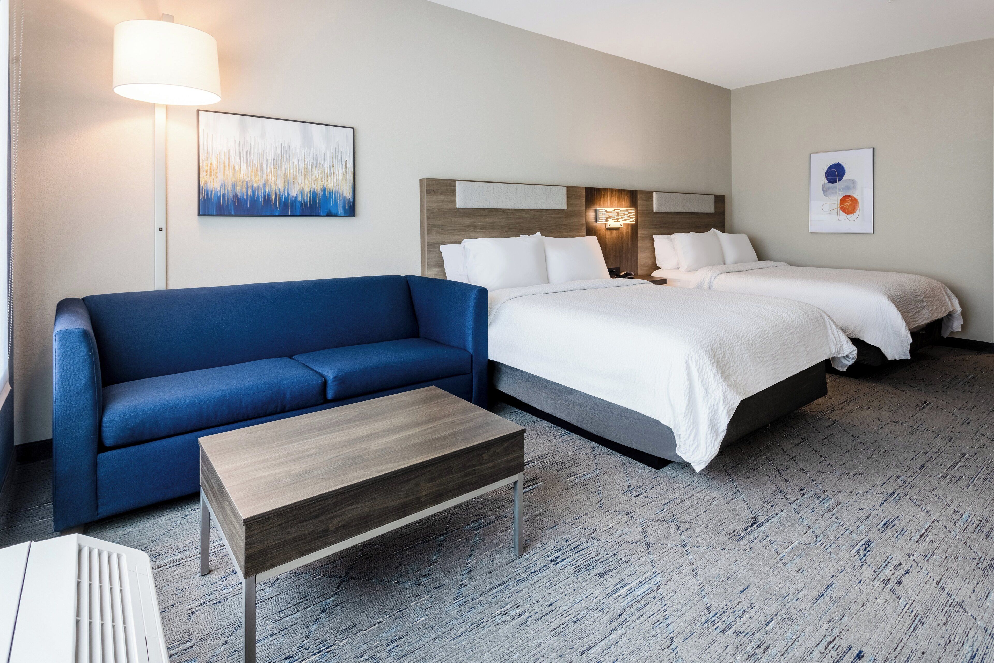 A hotel room with a blue sofa, a wooden coffee table, a large bed with white bedding, and two wall-mounted lamps.