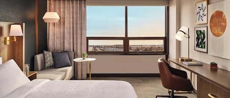 A hotel room with a large bed, a desk, a chair, a small table, and a view of the city through the window.