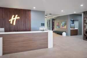 Un área de recepción moderna con un mostrador de madera, un gran letrero iluminado con la letra 'H', una zona de estar con un banco y una obra de arte colgada en la pared.