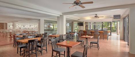 A spacious restaurant with wooden tables and chairs, ceiling fans, and large windows offering views of the outdoors.