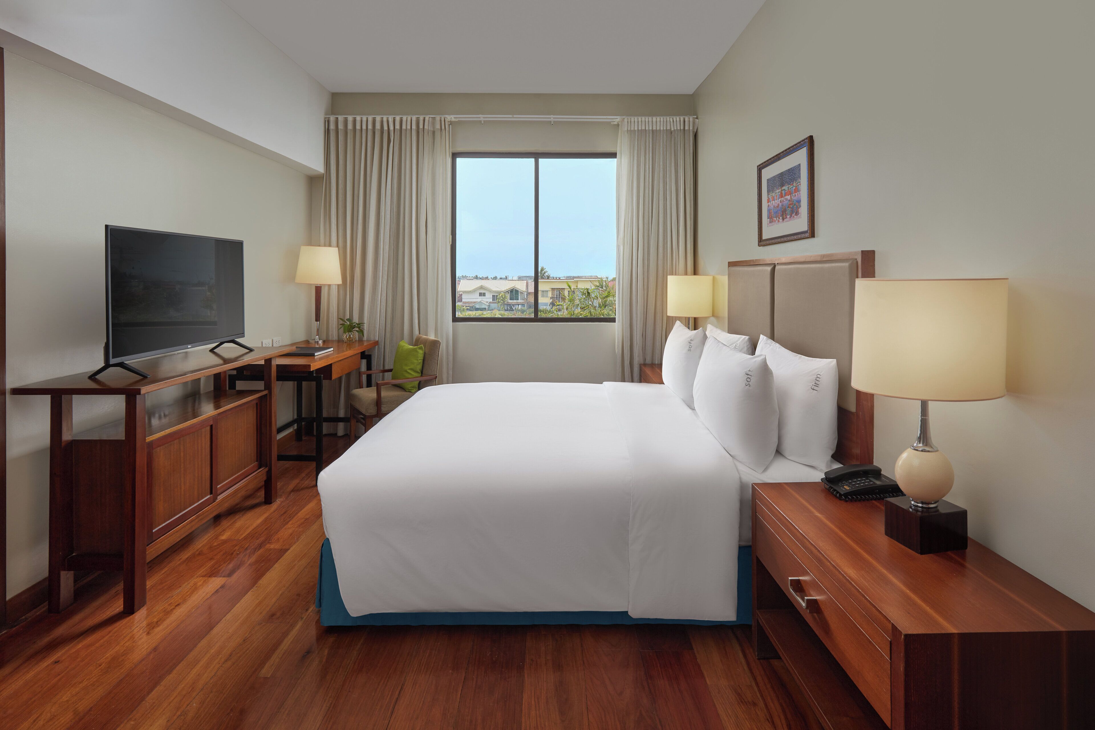A hotel room with a large bed, a TV, a desk with a lamp, and a window with a view of buildings.