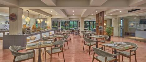 A spacious dining area with multiple tables and chairs, a central counter with food items, and large windows offering a view of greenery.