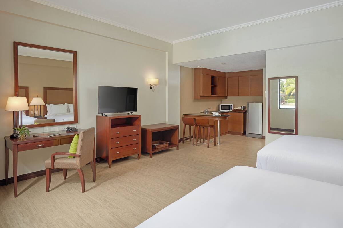 A hotel room with a desk, chair, TV, kitchenette, and two beds.