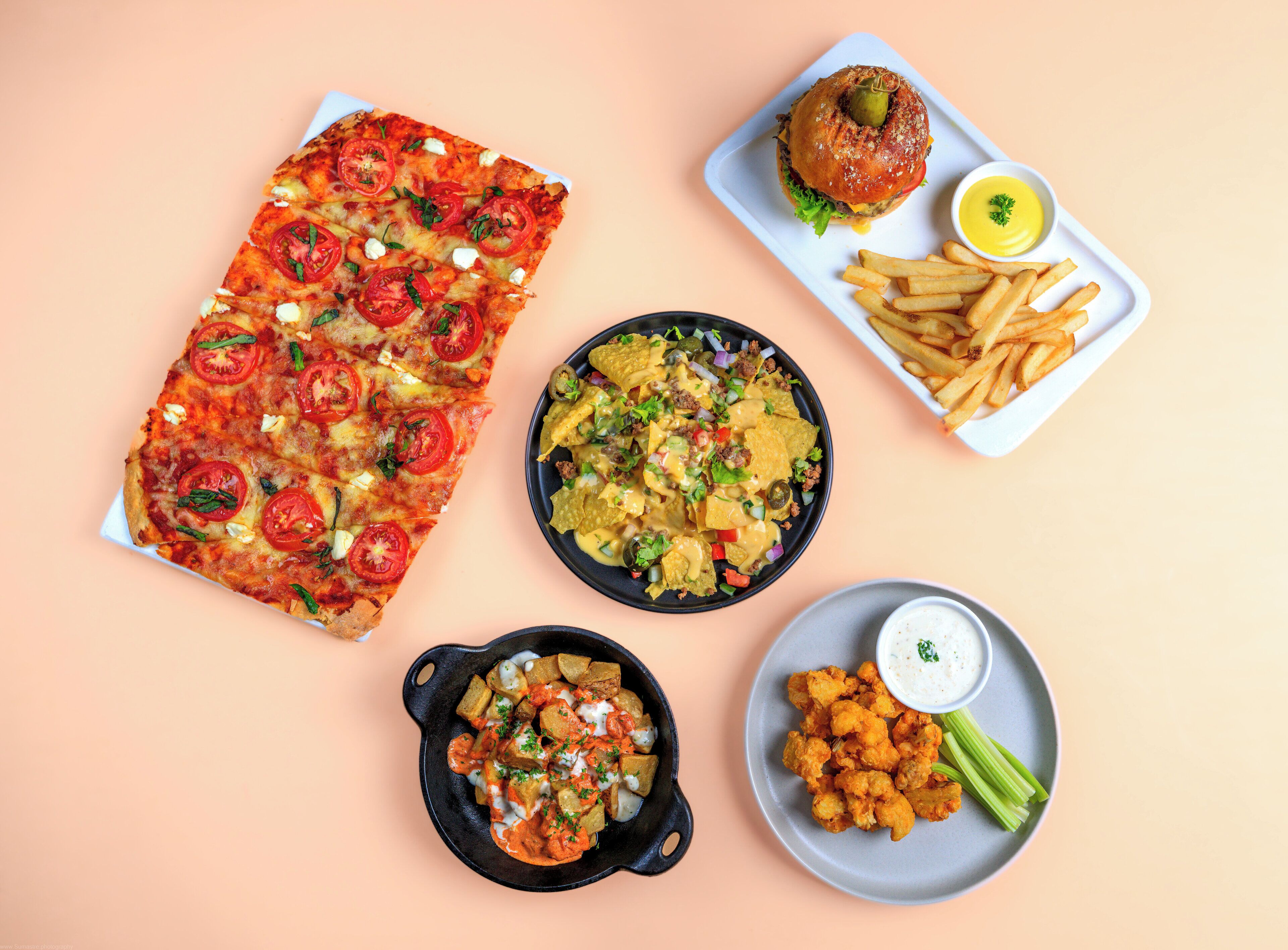 Verschiedene Gerichte wie Pizza, Nachos, ein Burger, Pommes und Chicken Wings, serviert auf unterschiedlichen Tellern.