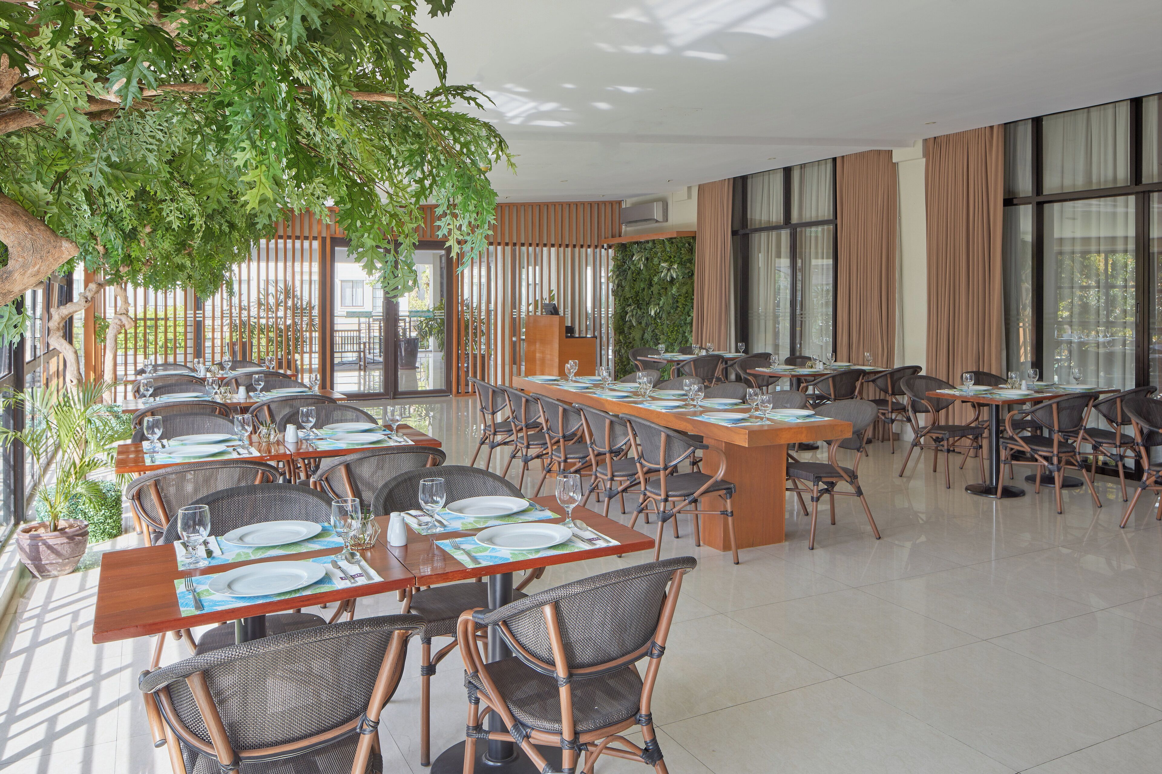 A spacious indoor dining area with multiple tables and chairs, large windows, and indoor plants.