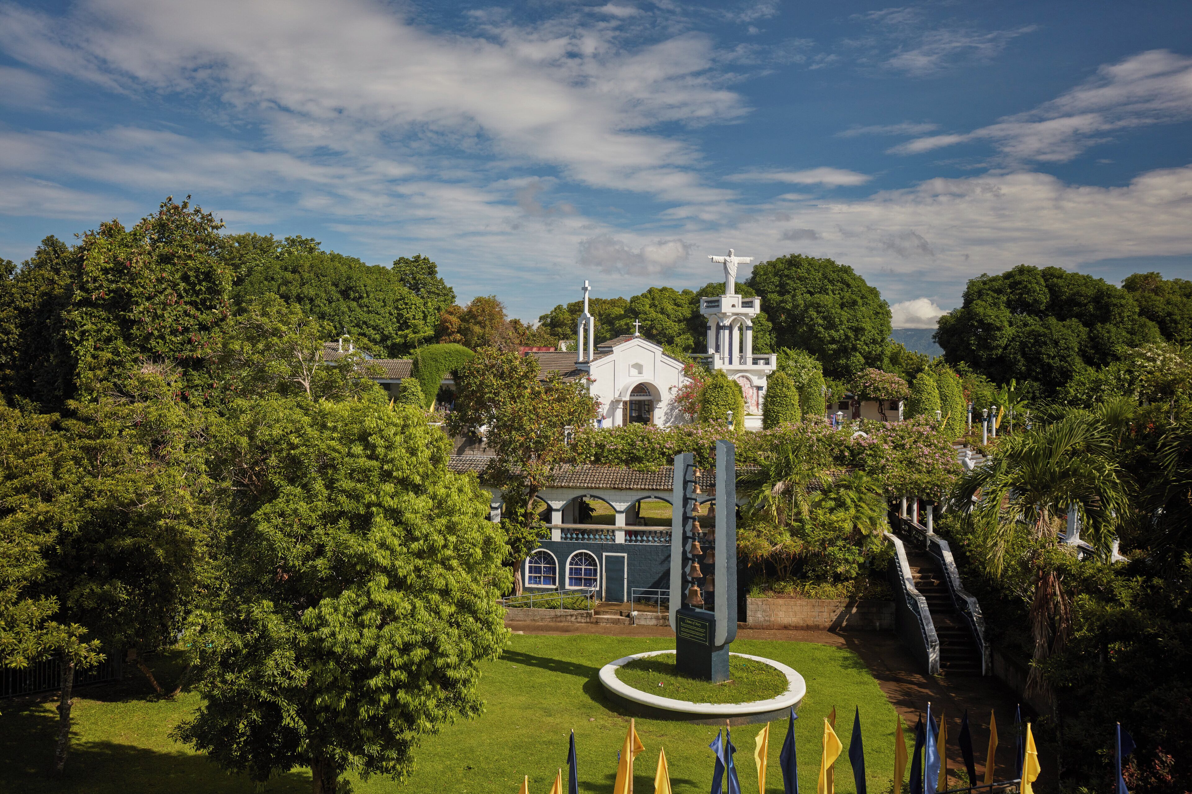 Una iglesia con campanario y estatua, rodeada de abundante vegetación y un monumento circular.