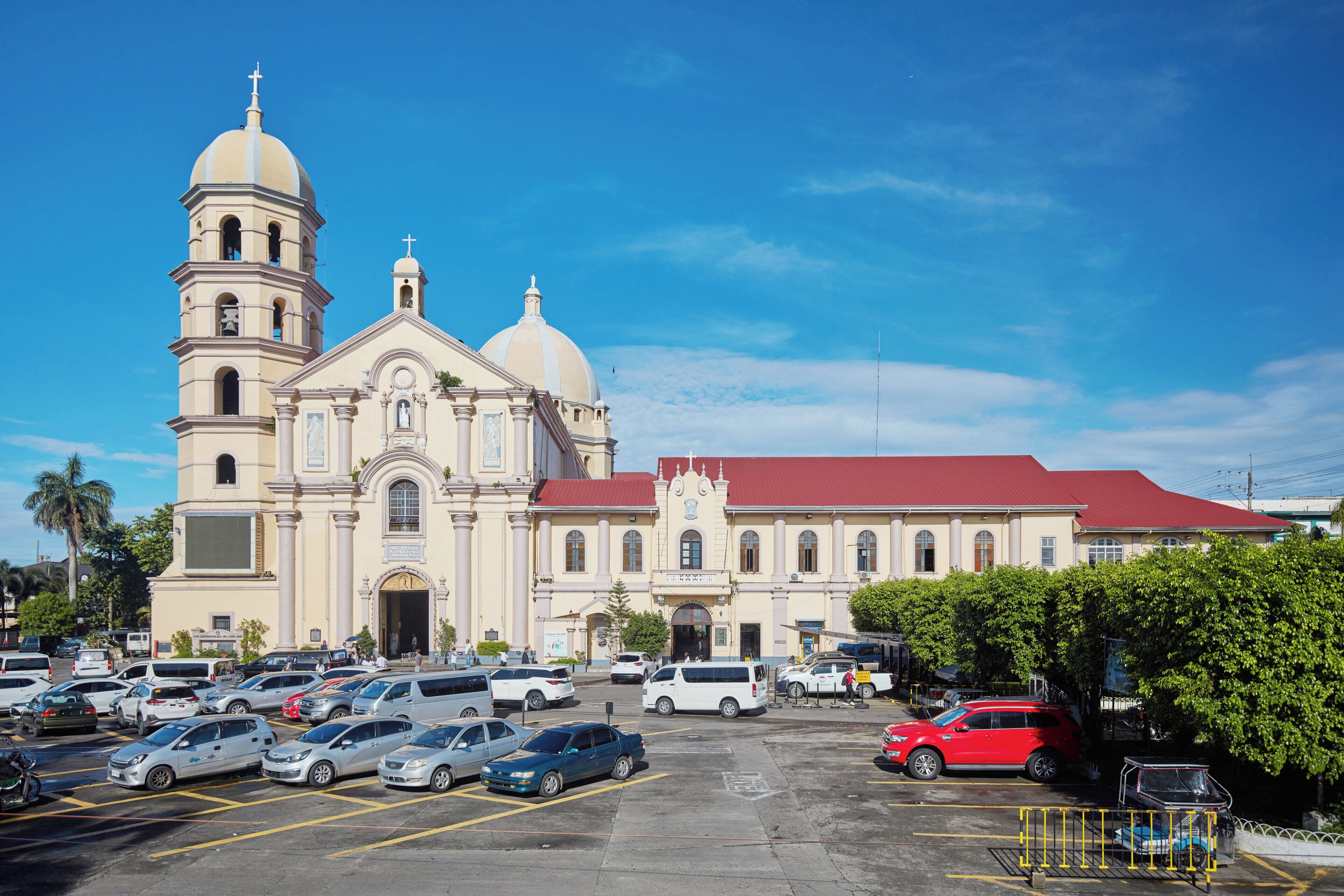 A large church with two domes and a bell tower, surrounded by a parking lot with various cars.