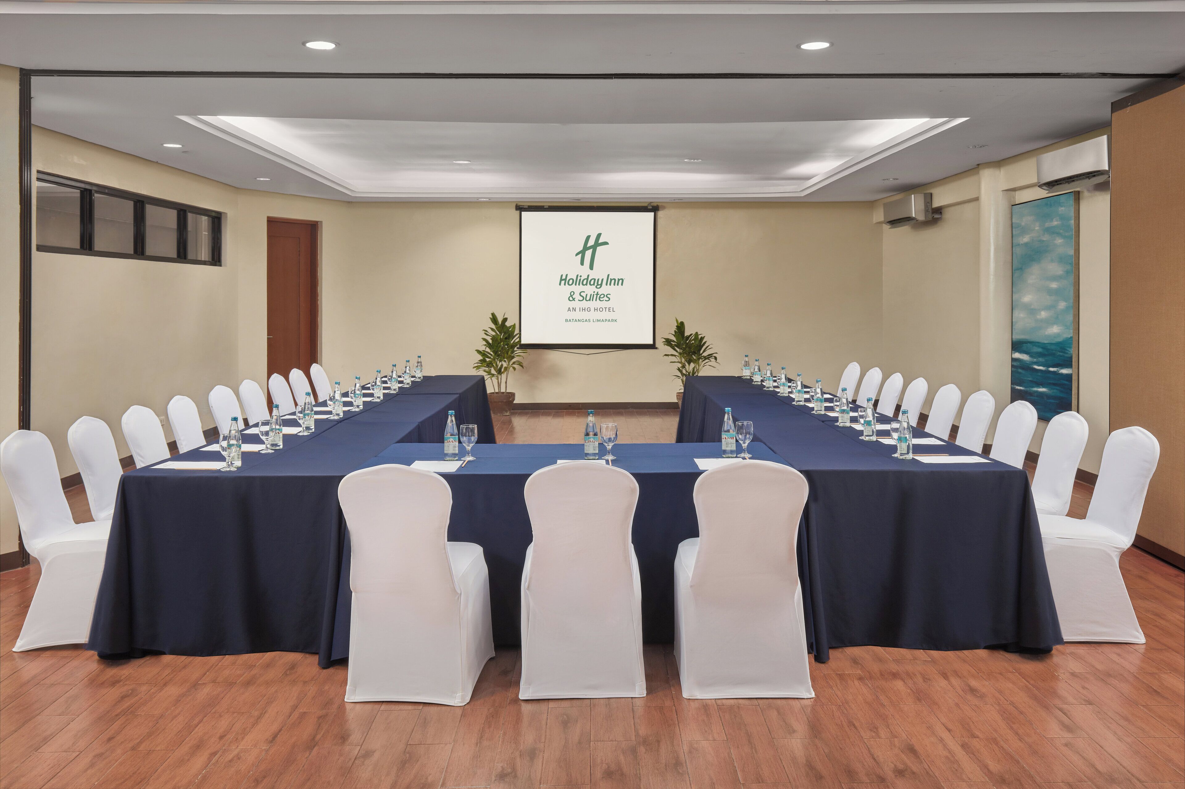 A conference room with a long table, white chairs, and a projector screen displaying a hotel name.