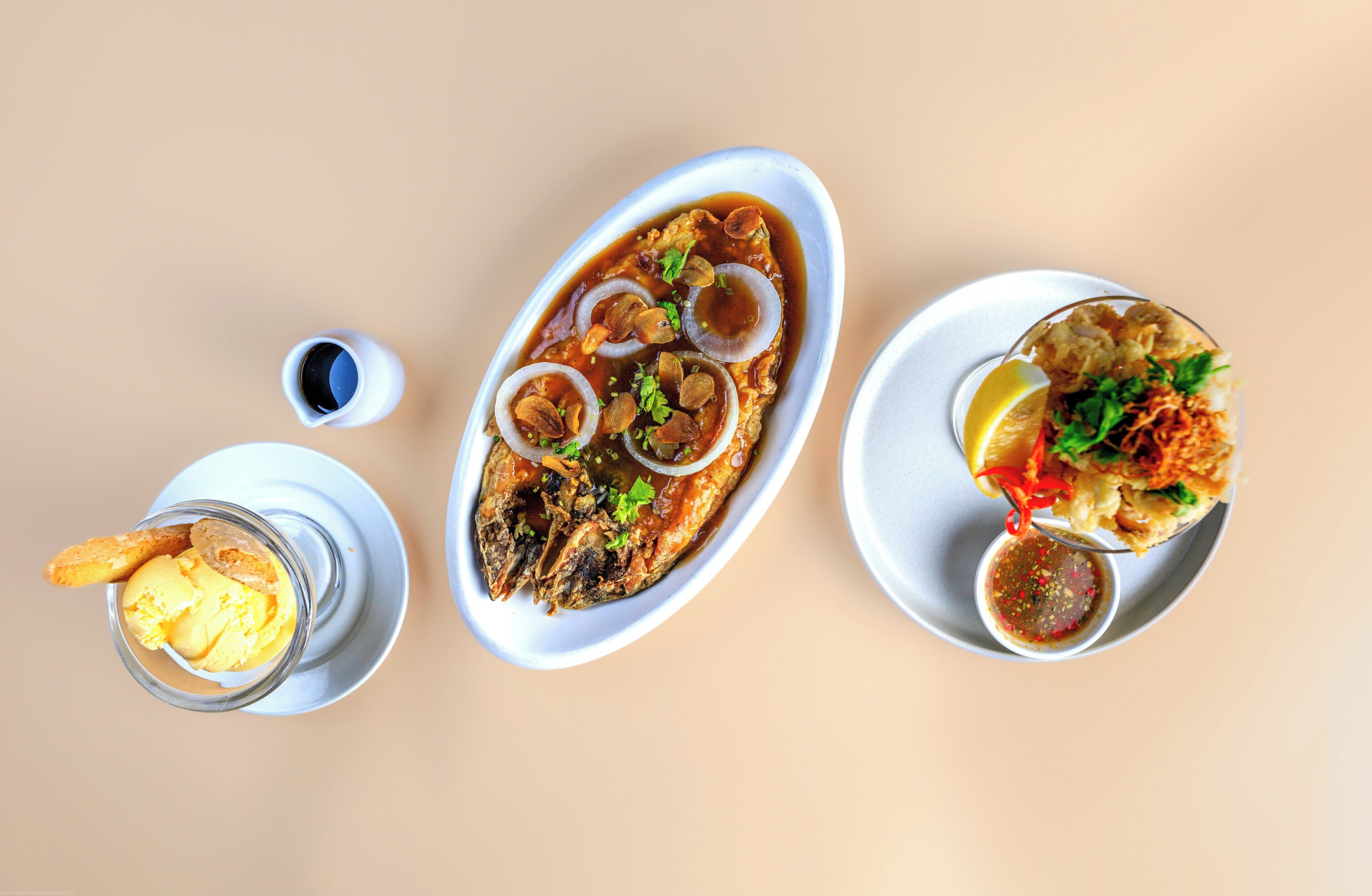 A table with three dishes: a fish dish with onions and herbs, a bowl of soup with a lemon slice, and a plate with vegetables and a sauce.