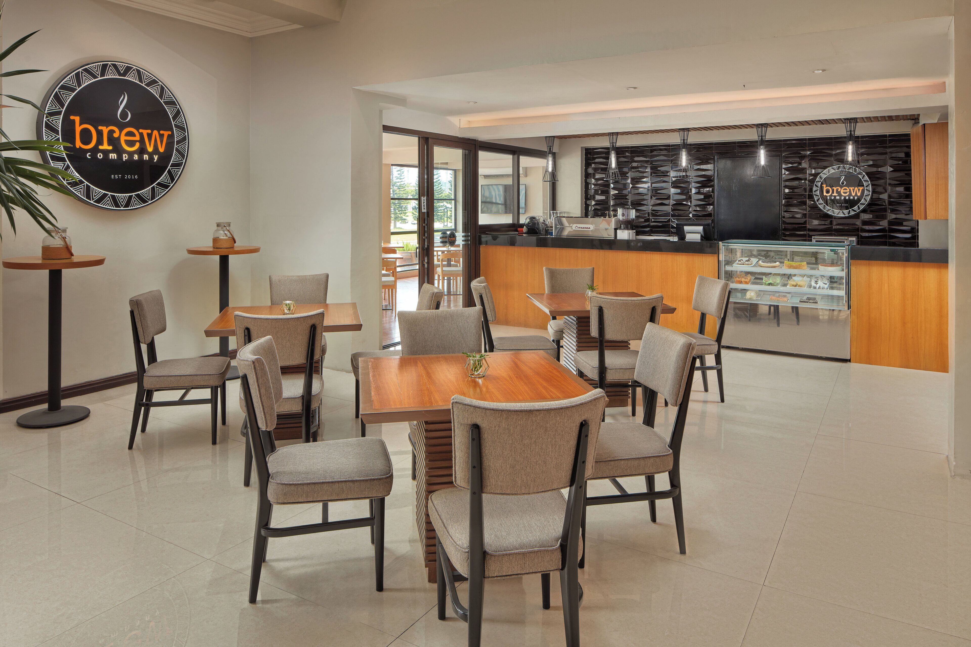 A modern cafe with a central wooden table surrounded by chairs, a coffee bar with a display case, and a logo on the wall.