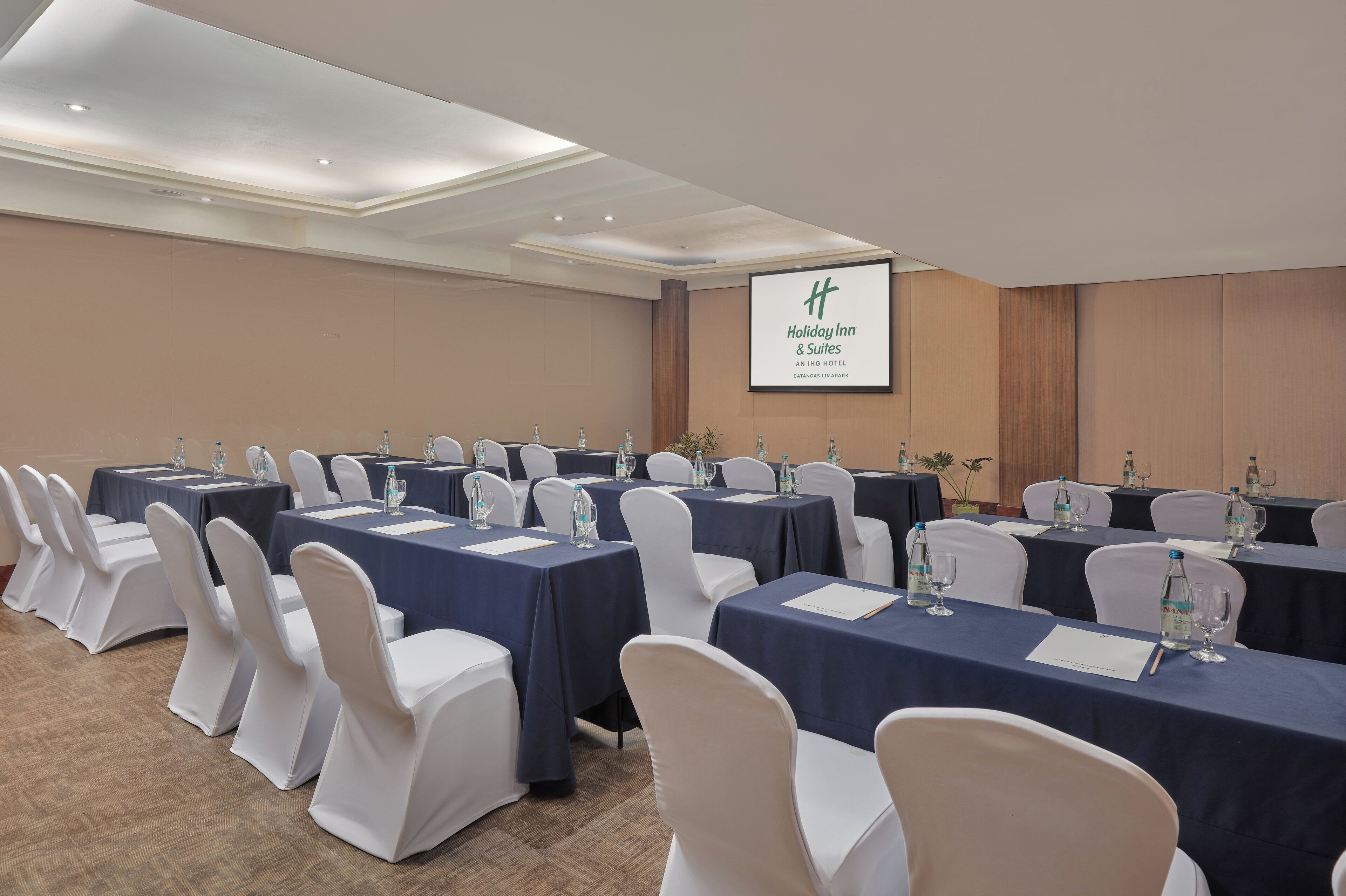 A conference room with tables set for a meeting, white chairs, and a screen displaying the Holiday Inn & Suites logo.