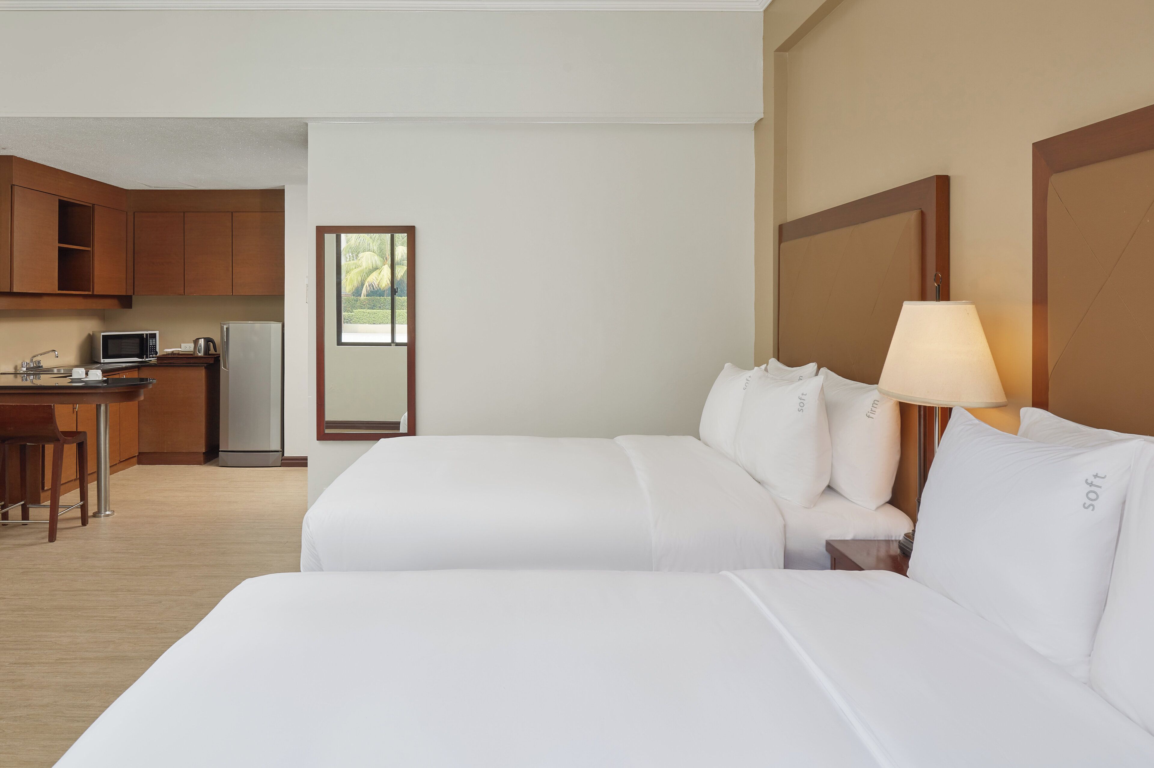 A hotel room with a kitchenette, two beds, a nightstand with a lamp, and a view of greenery through a window.