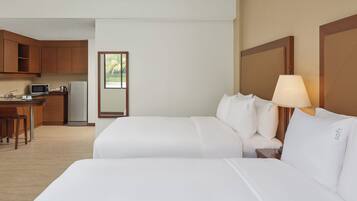 A hotel room with a kitchenette, two beds, a nightstand with a lamp, and a view of greenery through a window.