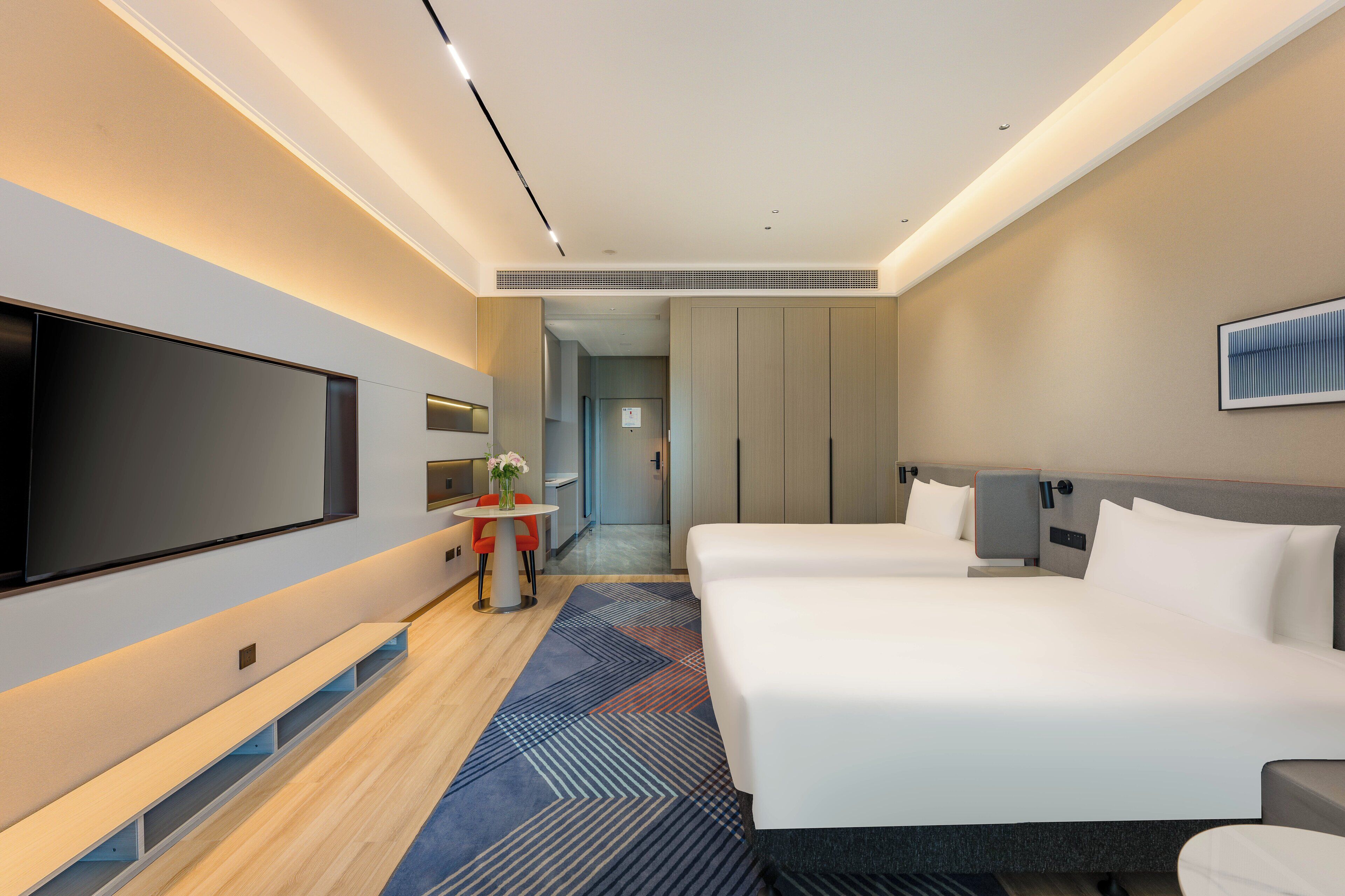 A hotel room with a large flat-screen TV, two beds, a small table with a vase, and a hallway leading to a bathroom.
