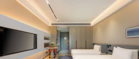 A hotel room with a large flat-screen TV, two beds, a small table with a vase, and a hallway leading to a bathroom.