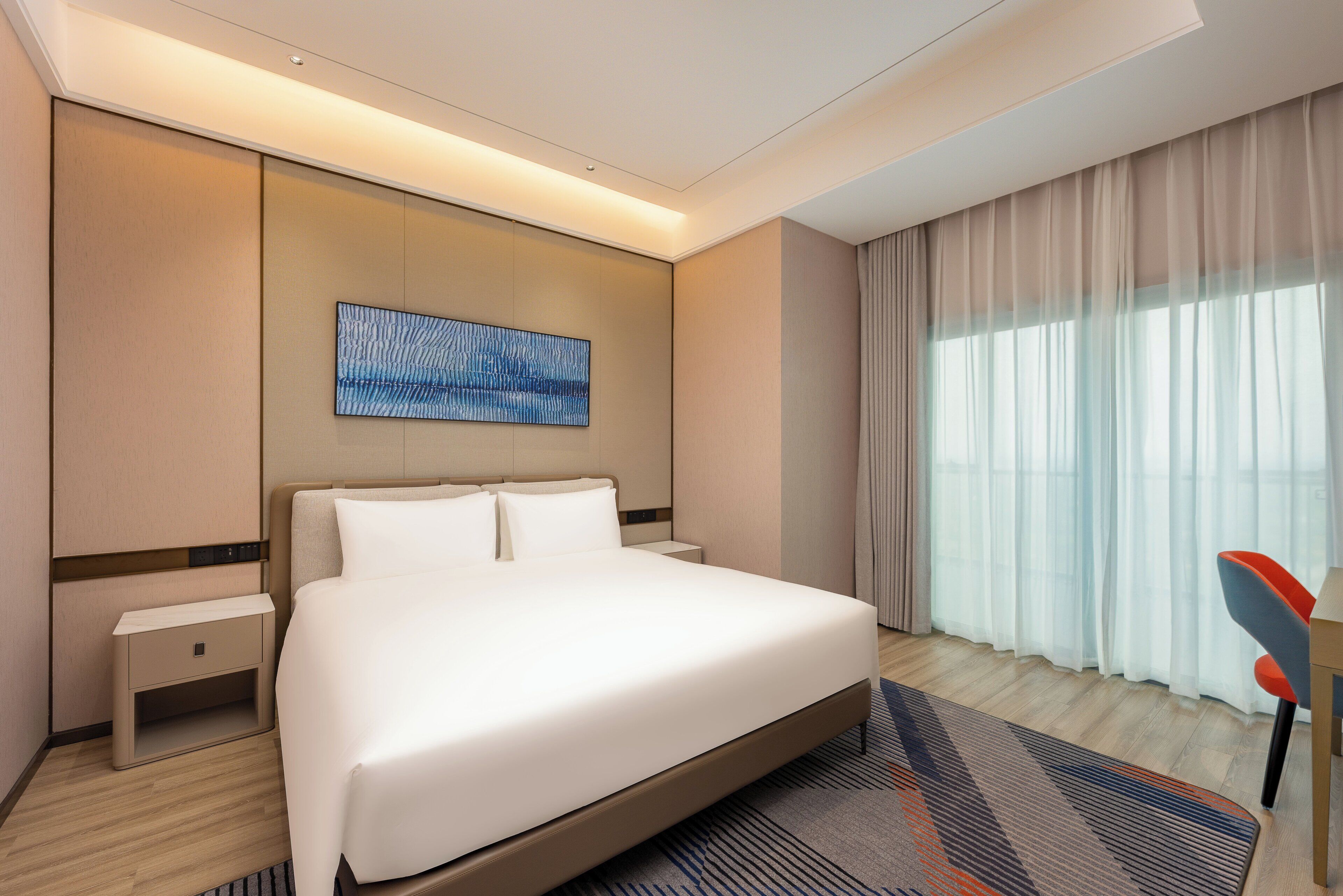 Una habitación de hotel moderna con una cama grande, una mesita de noche, una obra de arte en la pared y una ventana con cortinas translúcidas.