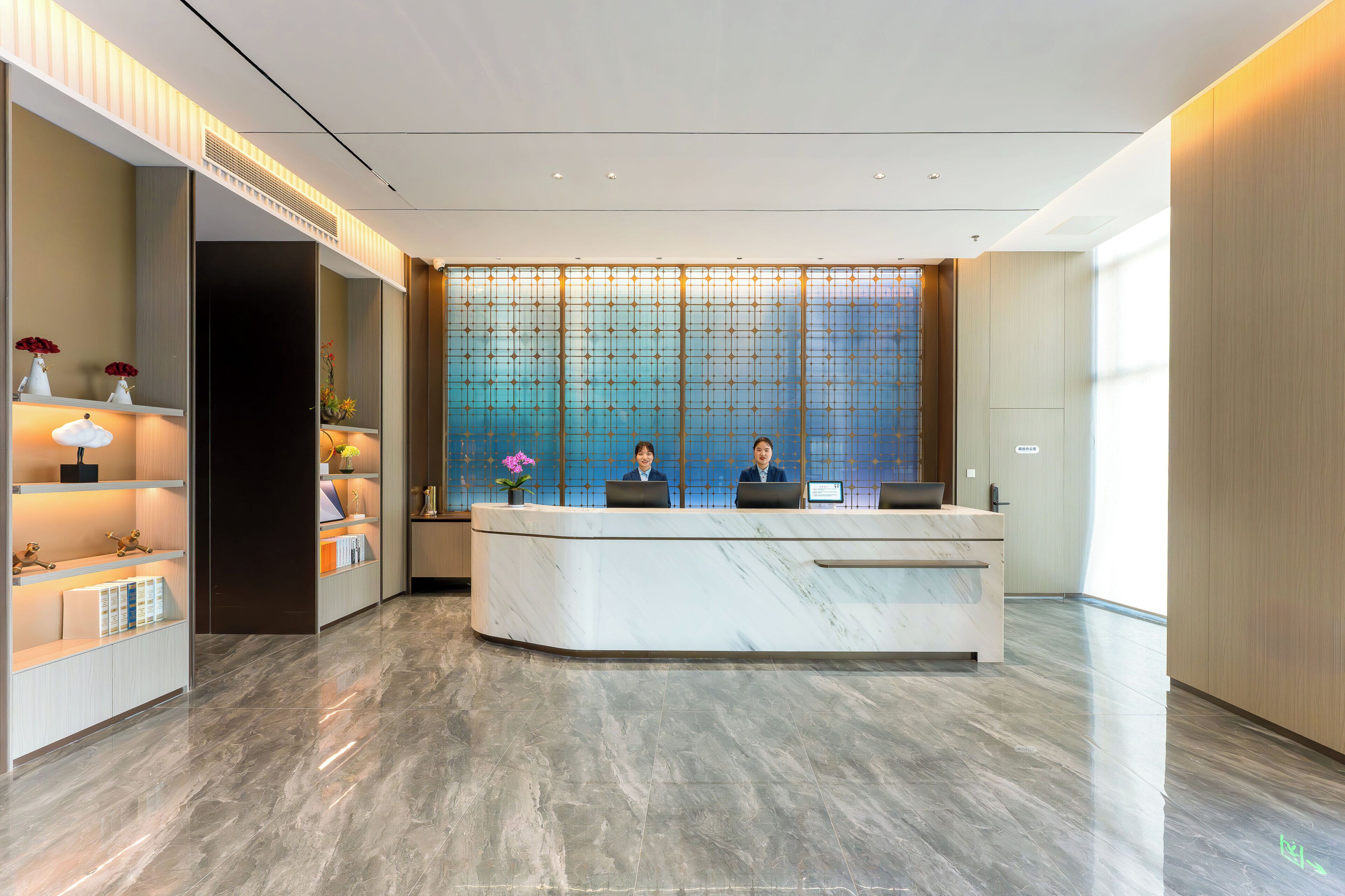 A reception desk with two staff members, a decorative wall with a geometric pattern, and a well-lit interior.